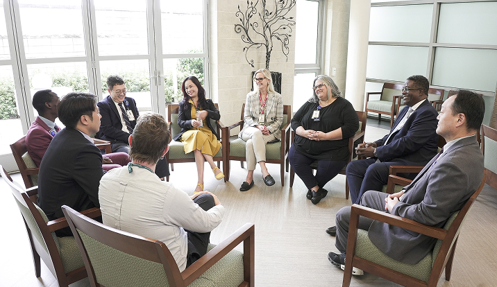 Group of colleagues in business attire seated in a circle, having a discussion in a bright office lobby.